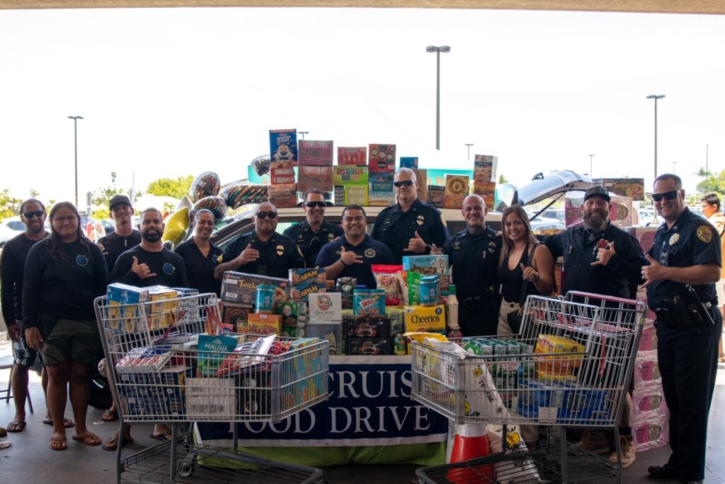 Volunteers and police officers in front of police car holding donated food during food drive.
