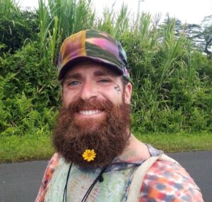 Kelly Gravitt is described as Caucasian, 6 feet tall, 180 pounds, with brown hair, blue eyes, and a beard. He also has several tattoos on his face and neck areas.