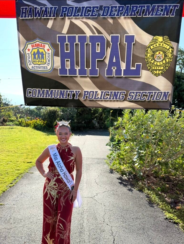 Miss Aloha Hawaii Scarlette Burnham-Rosario wearing a crown standing in front of a police banner.
