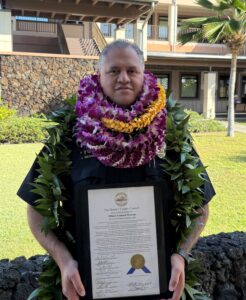 Officer Leonard Warren wearing several lei holding Haweo award outside Kona County building.