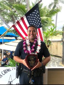 Kona Patrol Officer Michael Gusman in uniform and wearing lei around his neck holding his award for Kona Crime Prevention Committee’s Officer of the Month for October 2025.