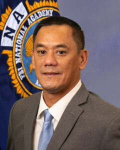 Captain Levon Stevens in a suit in tie posing by FBI National Academy flag.
